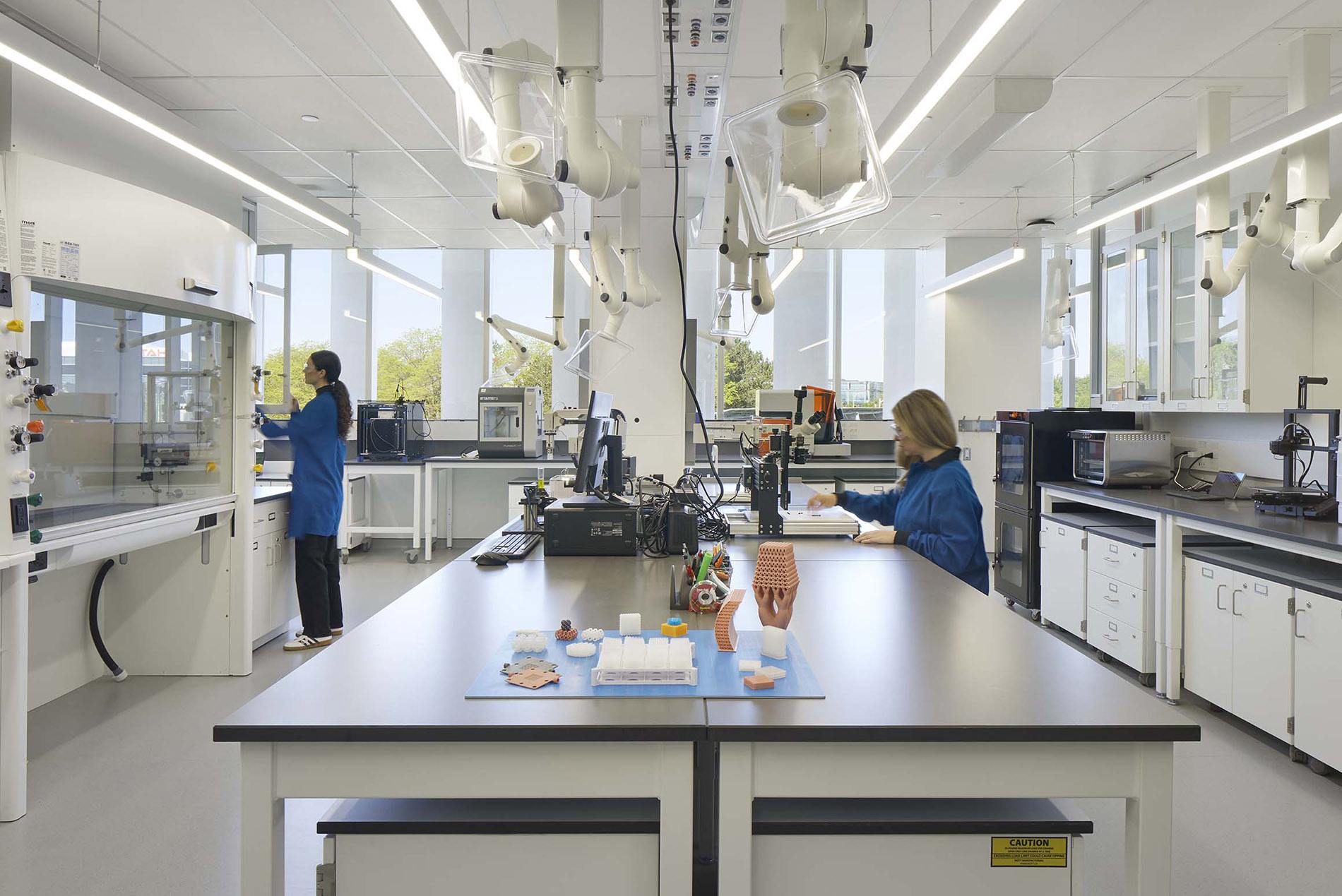 Two technicians in blue lab coats working at a fume-hood laboratory with overhead extraction arms
