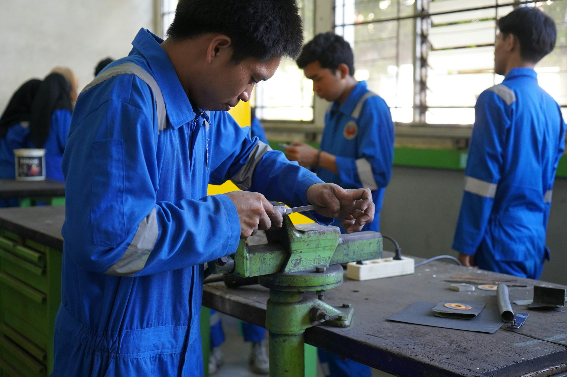 Vocational trainees in blue coveralls filing metal at bench vices in a workshop