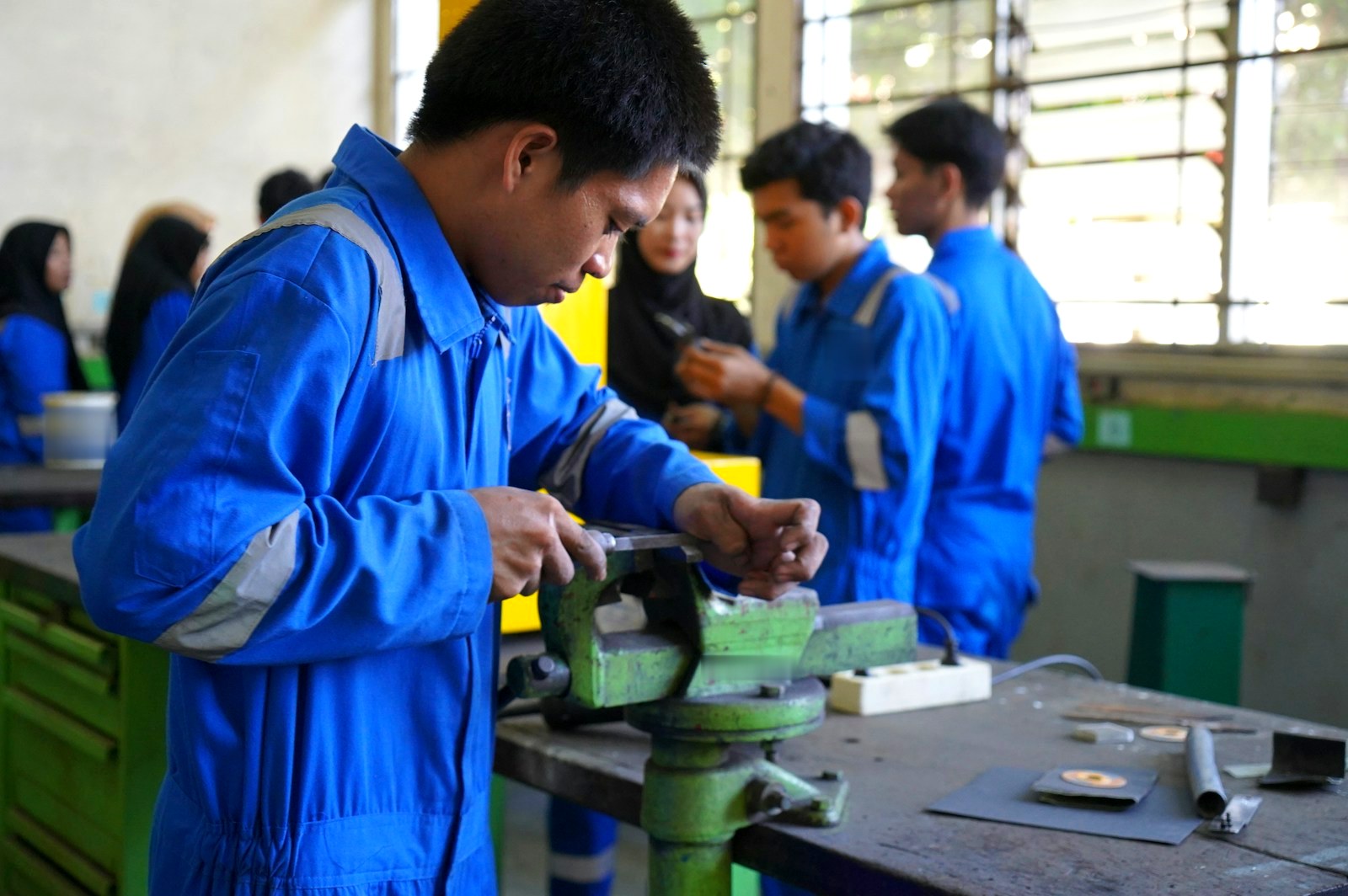Trainees in workshop coveralls filing a metal part at a bench vice during a practical class